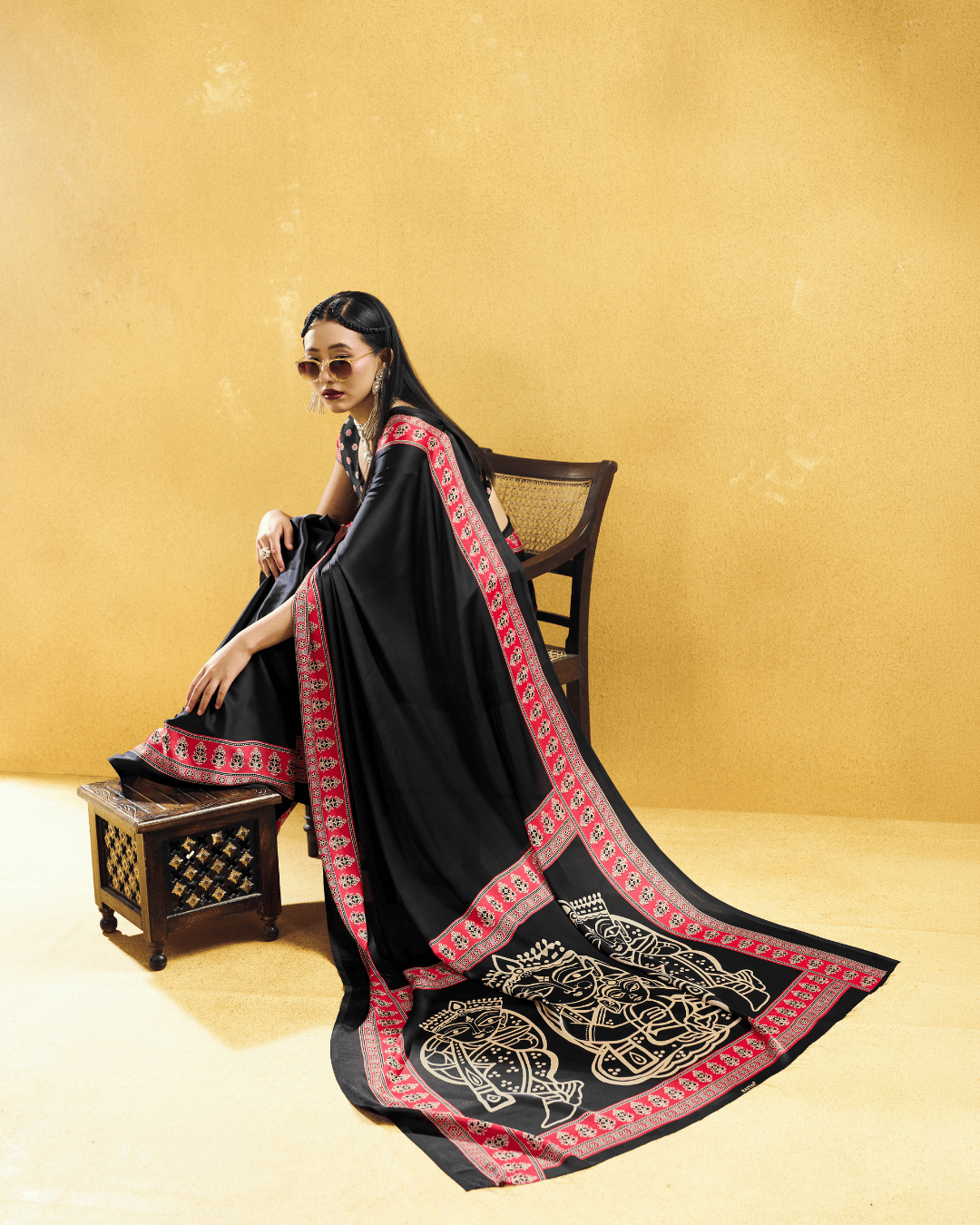Seated model showcasing a black pre-draped saree draped smoothly, highlighting the intricate red border details and large pallu artwork.