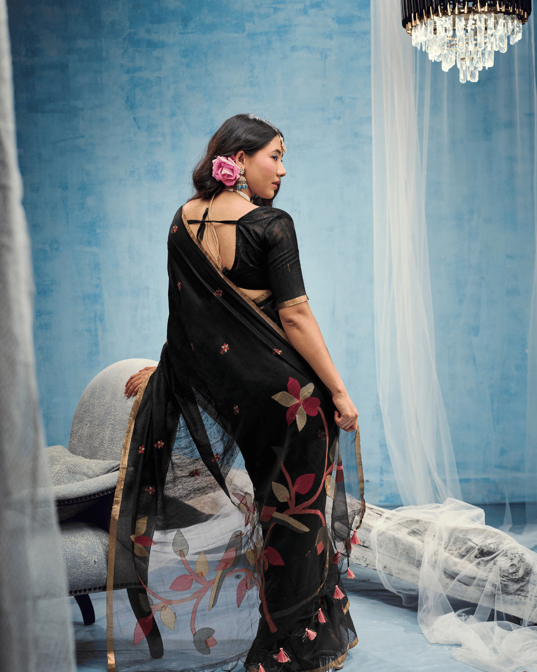 Back view highlighting the sheer black pallu with floral stems, oversized leaves, bird motifs, and small pink tassels hanging along the bottom edge.
