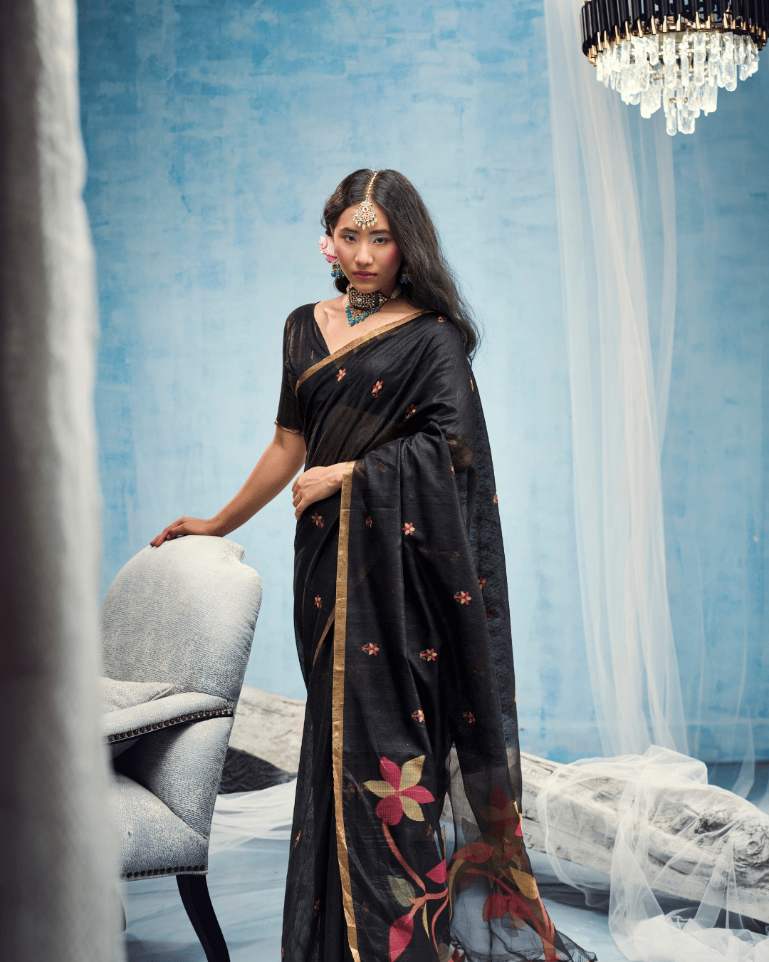 Model standing beside a grey armchair in the deep black ready to wear pocket saree, showing the front drape framed by a slim gold-toned border and accented with scattered floral motifs.