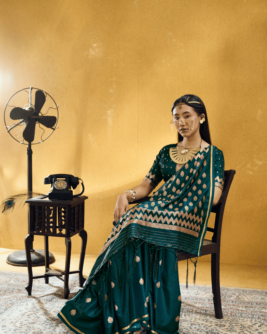Full-length portrait of a model seated on a dark wooden armchair wearing the Forest Green Chevron pre-stitched pocket saree, displaying pre-draped front pleats and gold piping along the edges.