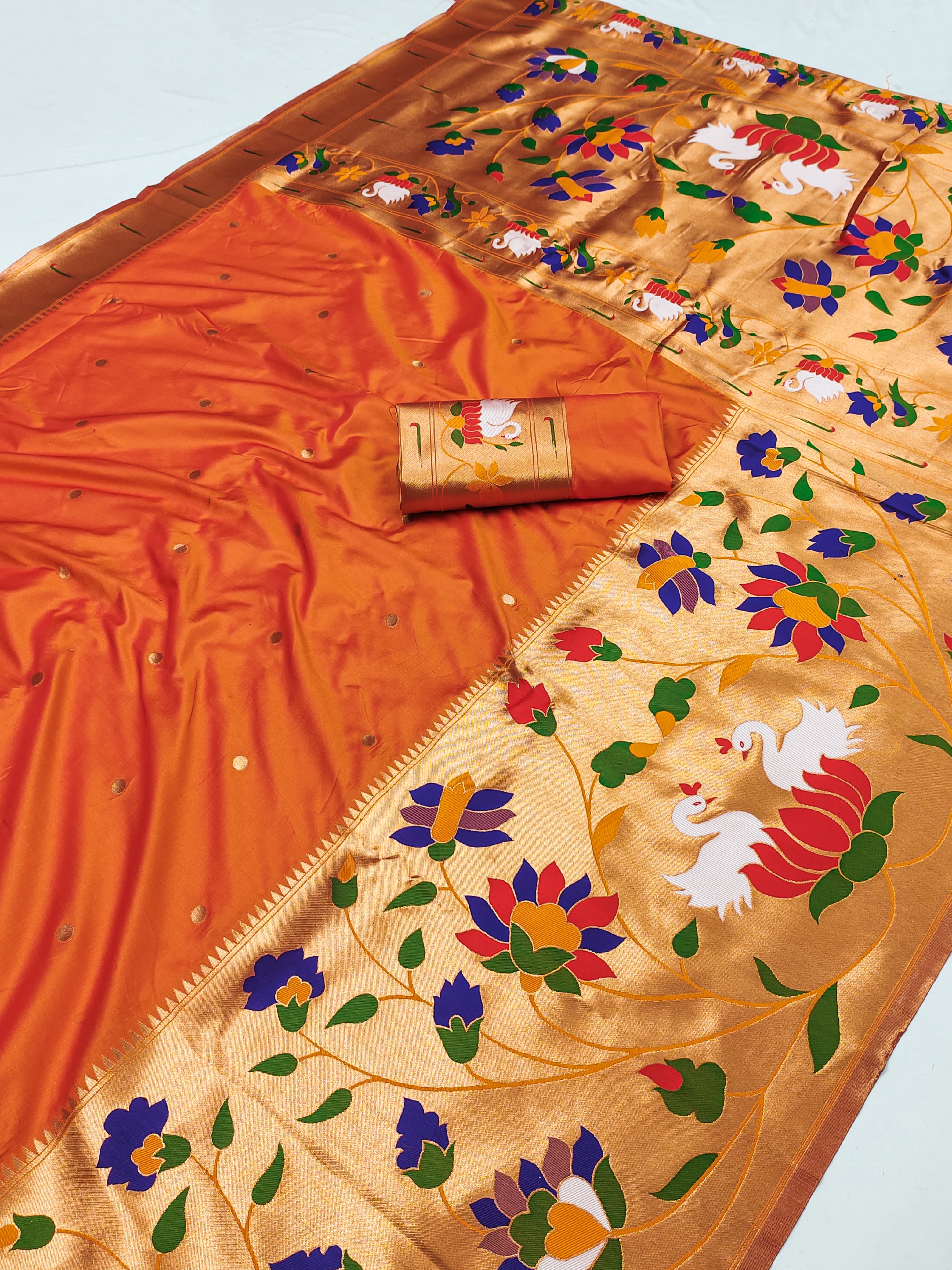 Flat lay of an orange Paithani silk saree featuring a golden-peach pallu woven with colorful flowers, green leafy vines, and white bird motifs.