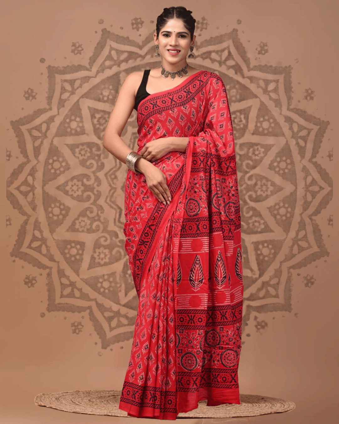 Coral-red Mul Cotton saree featuring small black and ivory leafy motifs, a geometric border, circular medallions, and large leaf prints on the pallu, separated by fine horizontal stripes.