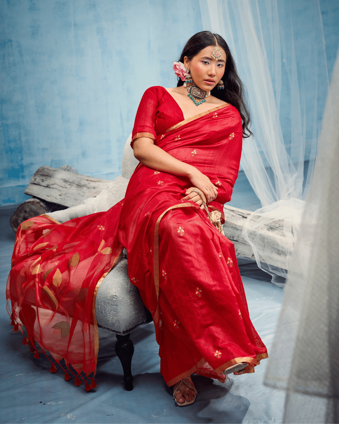 Model seated on a chair in the vibrant red cotton ready to wear pocket saree, showing the spread of scattered small floral motifs across the pleated drape and a slim golden border.