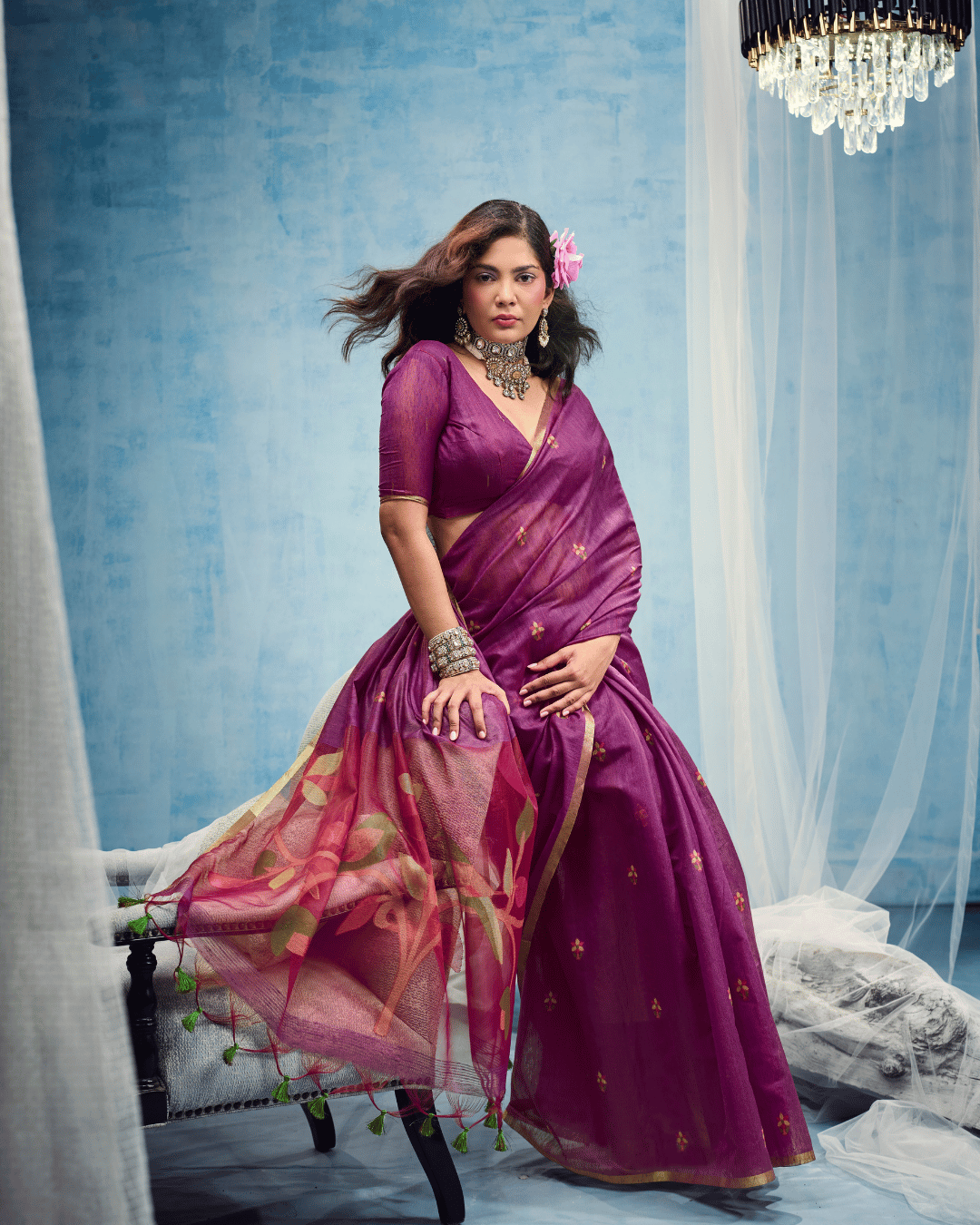 Model seated on an upholstered chair with the sheer pallu draped across the seat, highlighting the contrast between the understated deep-purple saree and the colorful botanical leaf artwork.