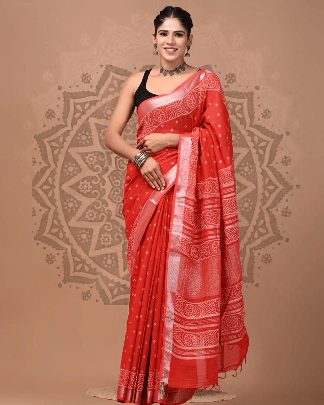 Coral-red linen saree with tiny pale diamond motifs, a pale pink-silver border, floral medallion and geometric pallu bands, horizontal stripes, and red tassels.