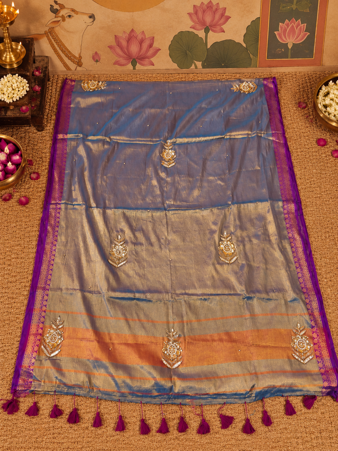 Flat-lay detail view of a soft silk tissue ready-to-wear pocket saree in a muted blue-grey tone, featuring silver and gold ornamental bootis, multi-colored striped pallu bands in rust and gold, a purple border, and deep purple tassels.