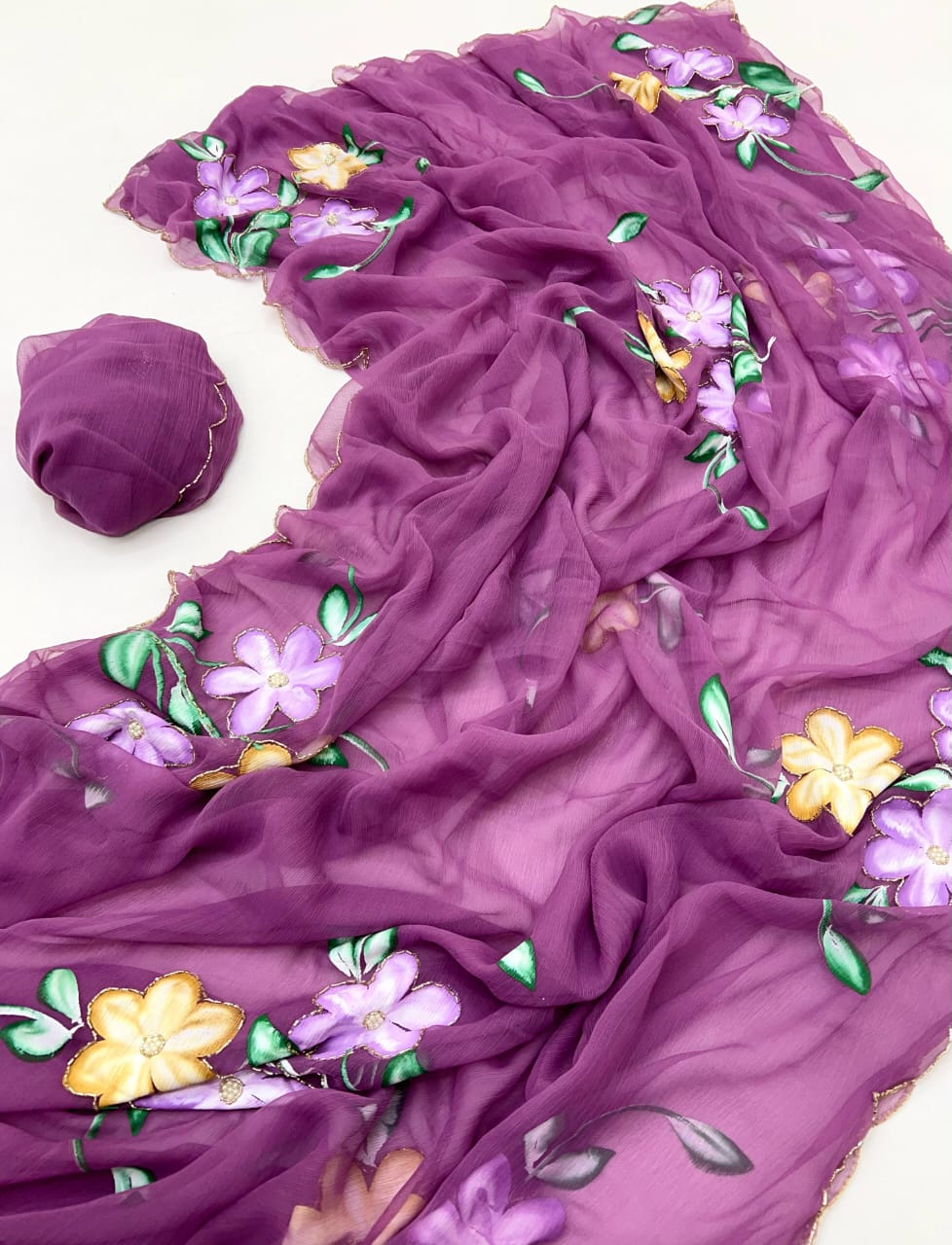 Flat-lay detail shot of the lightweight purple chiffon saree, showcasing large khatli handwork style lavender and yellow floral blooms, leafy green stems, and a fine gold-embroidered scalloped border.
