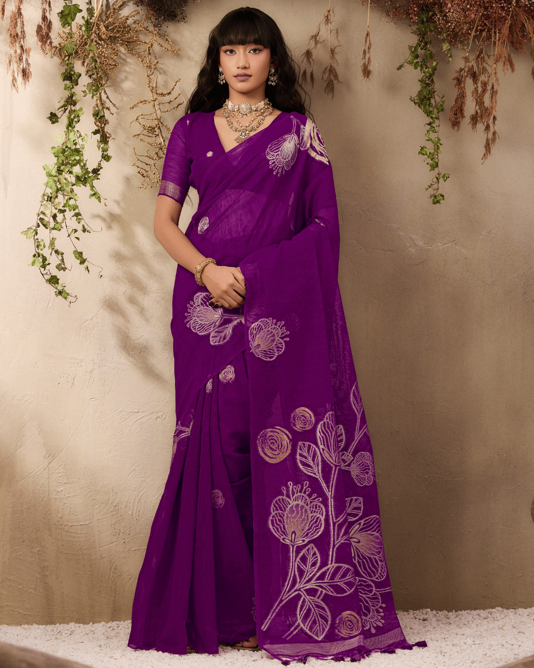 Full-length portrait of the model standing against a textured neutral backdrop framed with dried foliage, displaying the pre-draped purple saree with scattered circular gold body motifs and a statement open pallu featuring large rose and leaf artwork.