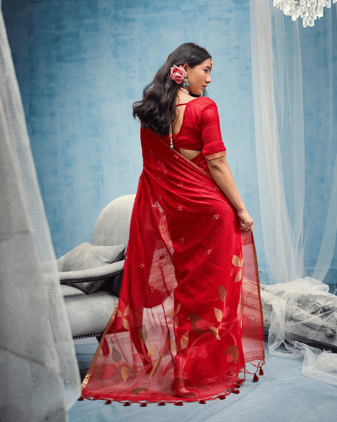 Back view of the model standing against a soft blue backdrop, showcasing the sheer red pallu decorated with large leaf motifs and small red tassels along the hem.   