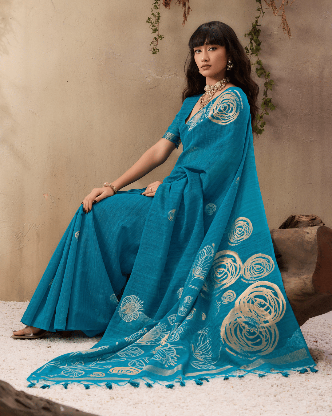 Full-length shot of the model seated beside a wooden log, displaying the pre-draped teal saree with its statement gold rose-and-leaf pallu and tassel ends.