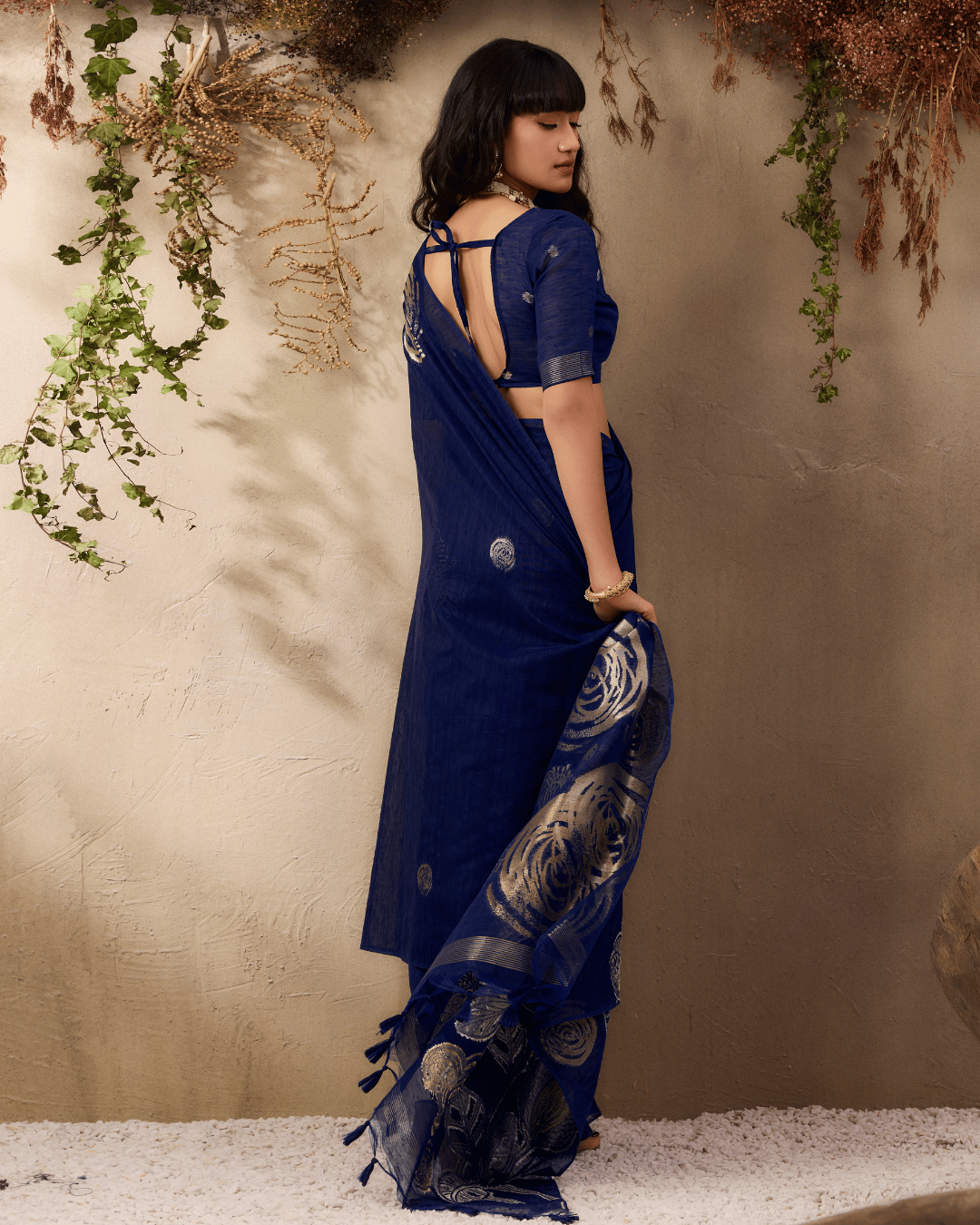 Back view of the model showcasing the deep navy blue saree drape, featuring subtle body motifs and the wide spread of the gold-outlined floral pallu.