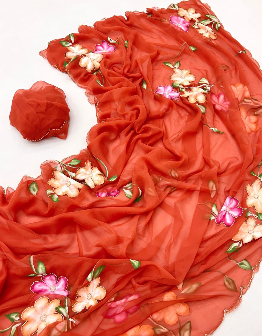 Flat-lay detail of a warm vermillion chiffon saree featuring handwork-style floral clusters in vibrant pink and soft yellow with leafy green stems, displayed with a delicate scalloped edge.